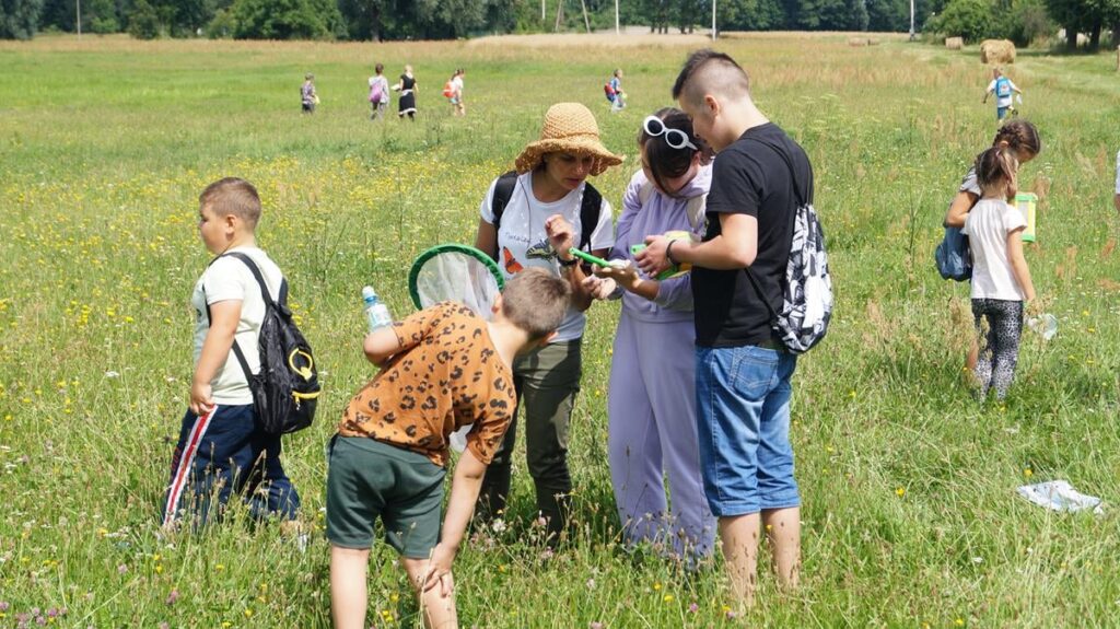 Grupka ludzi na łące pochyla się nad  znaleziskiem przyrodniczym. 