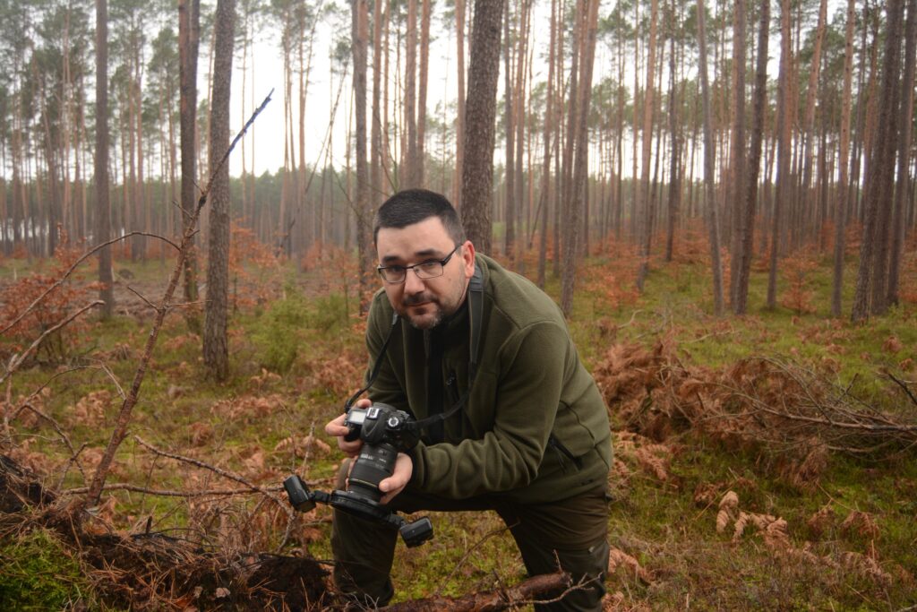 mężczyzna w okularach pochylony z aparatem w ręku stoi w rzadkim lesie