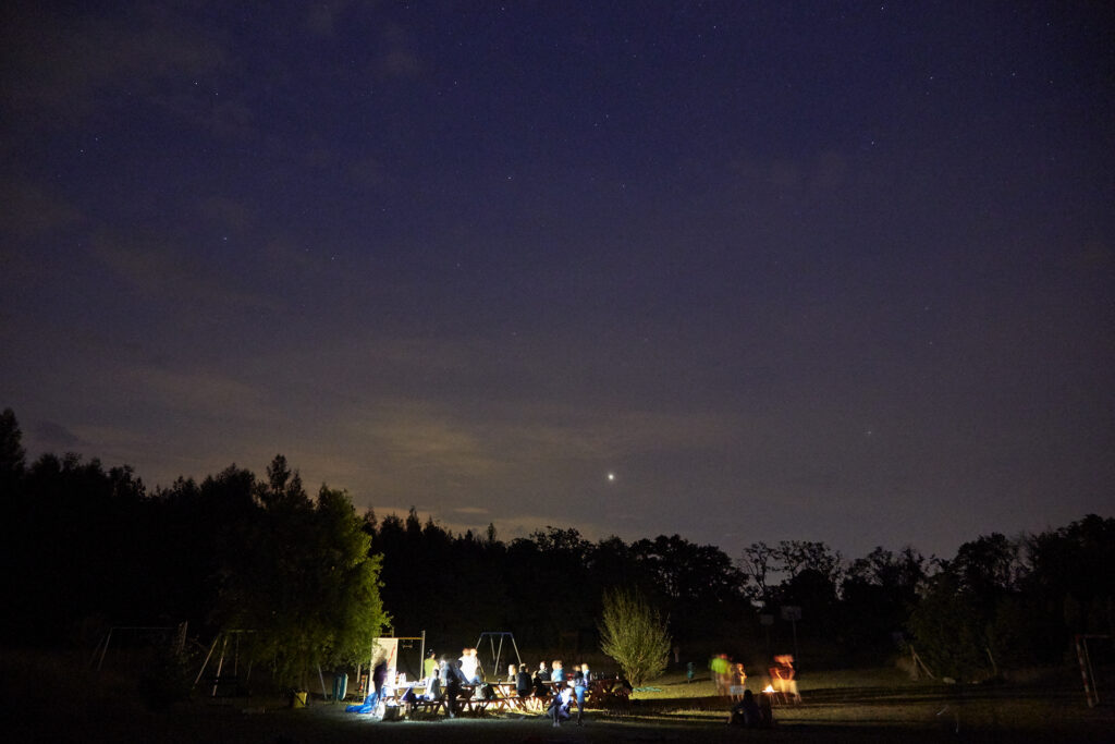Nocne zdjęcie przedstawiające ludzi obserwujących spotkanie przy świetle. Widać także nocne niebo, świecącą się planetę oraz sylwetki drzew. 