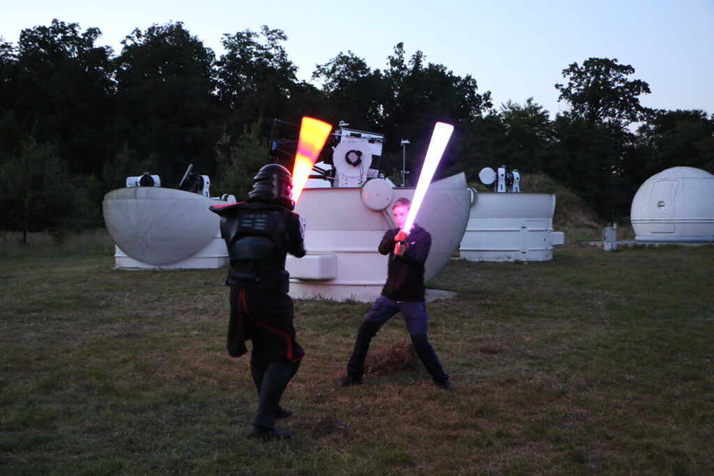 Dwie osoby walczą na miecze świetlne. Jedna z nich jest przebrana  w strój postaci z Gwiezdnych Wojen. 
